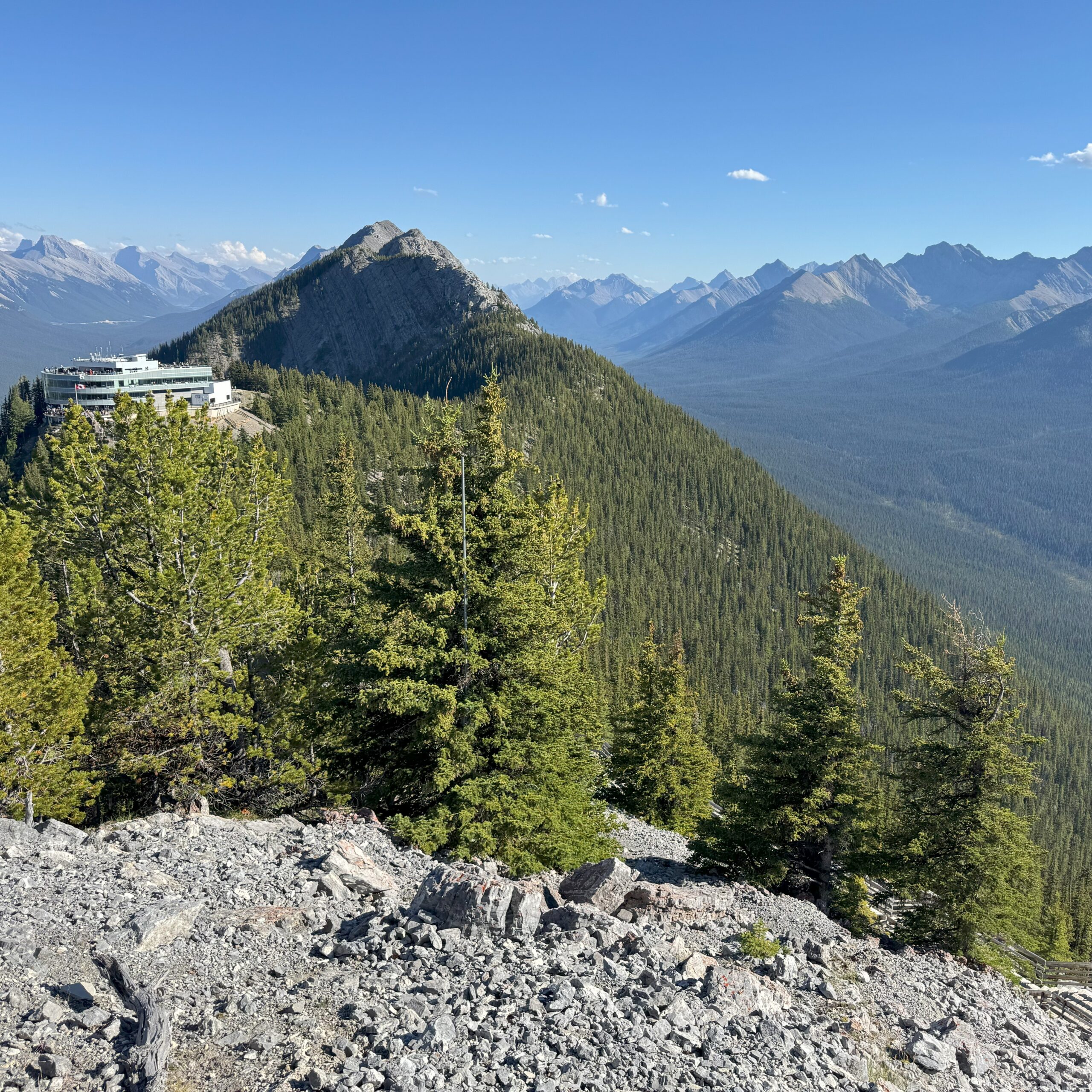 View from Sulphur Mountain in Banff, showing the Banff Gondola station on the left and a forested mountain ridge stretching into the distance under a clear blue sky.