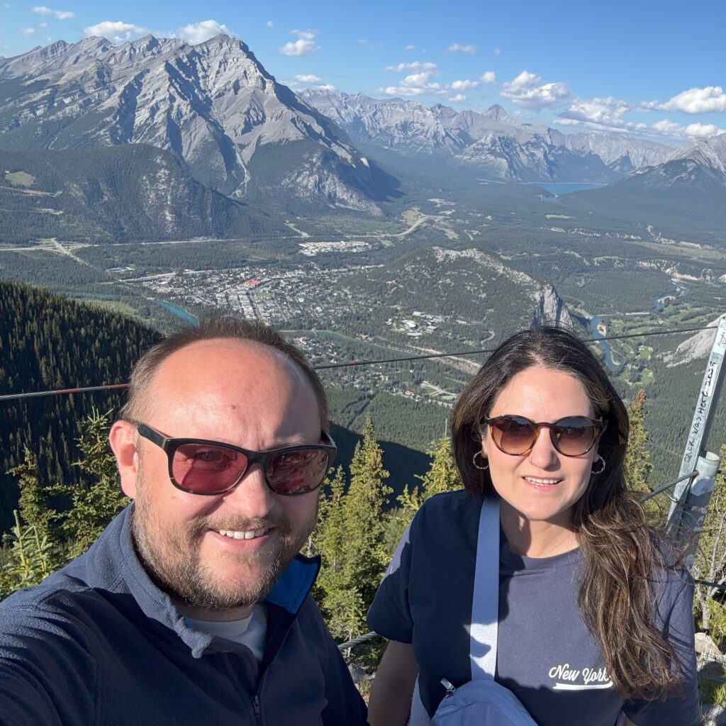 View from the Cosmic Ray Station Two people smiling in the foreground with sunglasses on, standing at a scenic mountain viewpoint. Behind them is a sweeping view of Banff town nestled among dense forests and towering Rocky Mountain peaks under a clear blue sky.