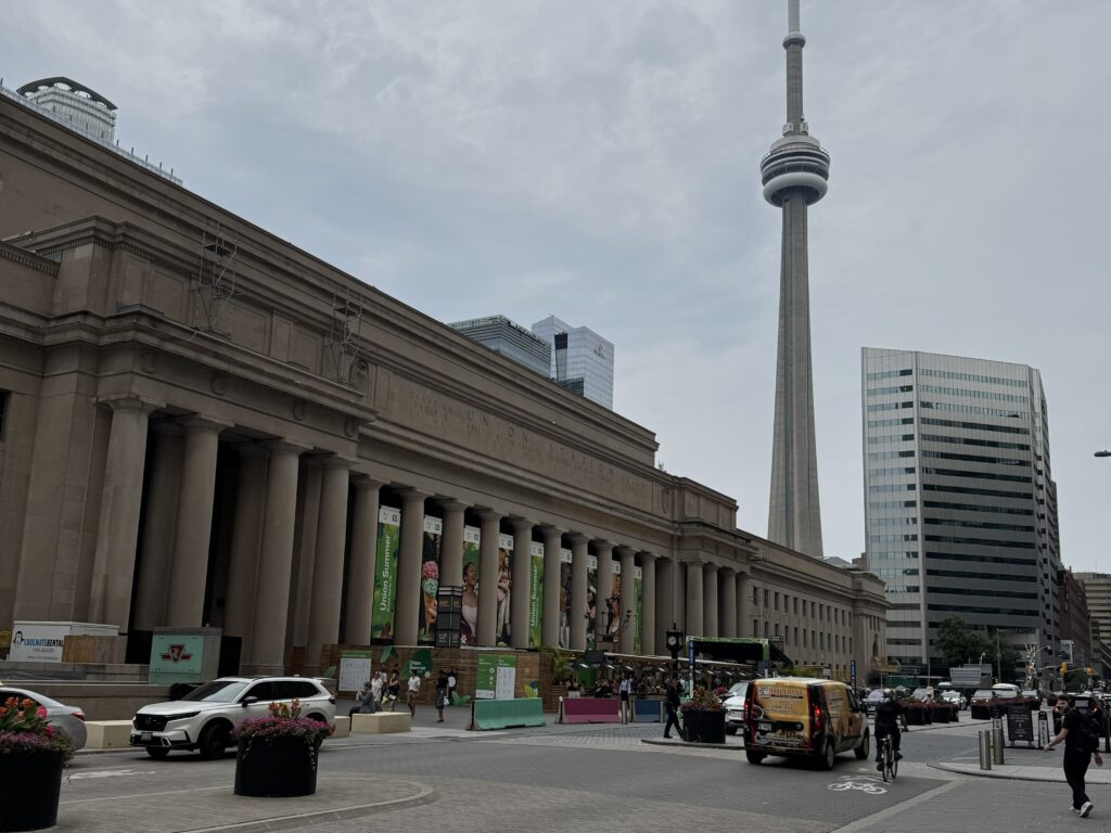 View of Toronto’s CN Tower rising behind Union Station on a cloudy day, with cars, cyclists, and pedestrians in the foreground.