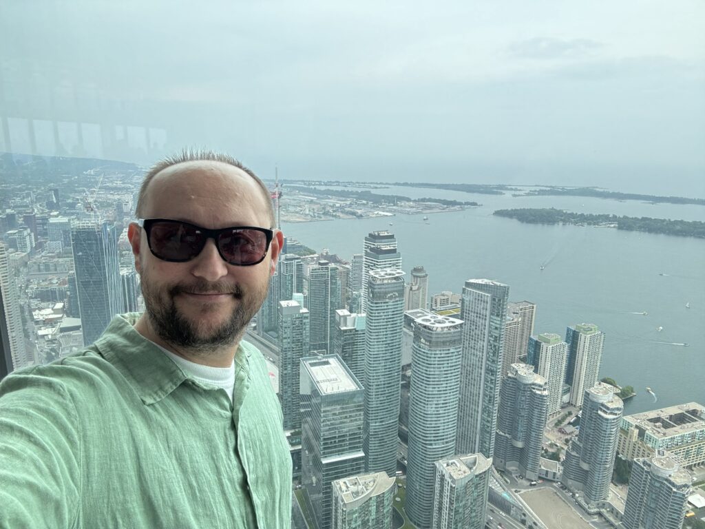 A person taking a selfie from the CN Tower observation deck with a panoramic view of downtown Toronto and Lake Ontario in the background.