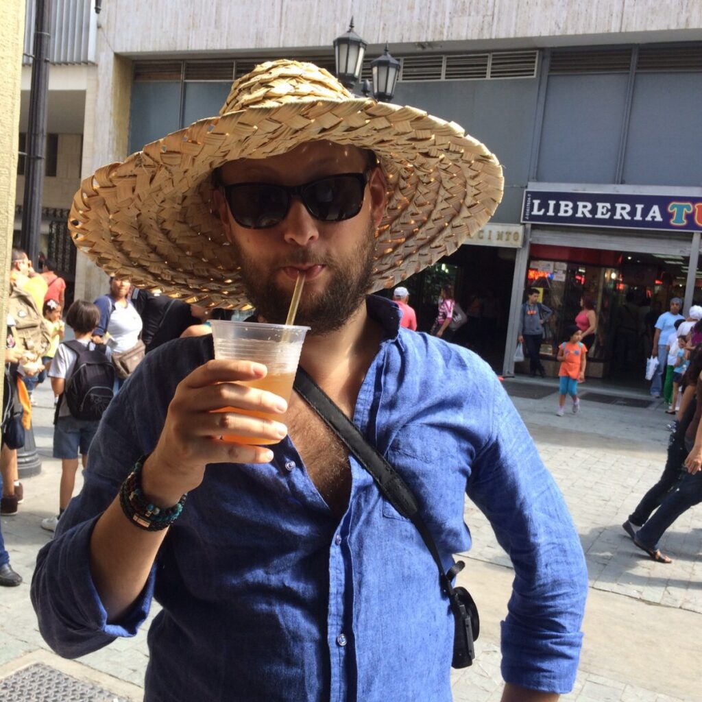 A person wearing a wide woven straw hat and sunglasses drinks a cold beverage from a plastic cup while standing on a busy street in Caracas, with people and storefronts visible in the background.