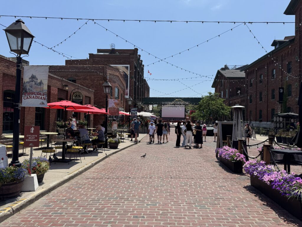 Visitors strolling along the cobblestone streets of Toronto’s Distillery District on a sunny day, surrounded by red-brick buildings, outdoor cafés with red umbrellas, and hanging string lights.