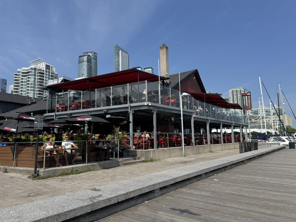 A waterfront restaurant with outdoor seating and red accents at Toronto’s Harbourfront, with modern high-rise buildings in the background under a clear blue sky.