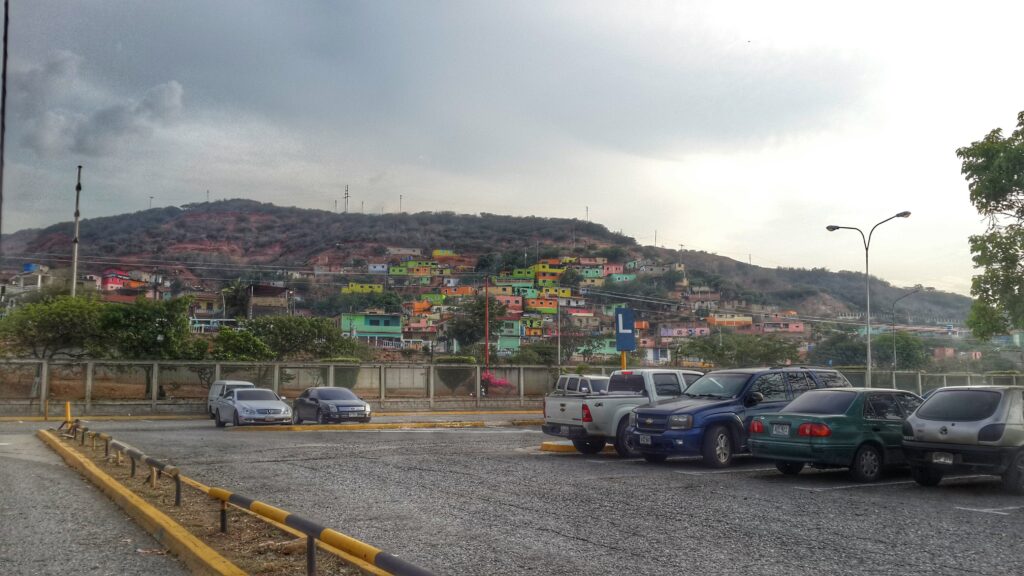 A view of colorful hillside houses near Caracas Airport, with parked cars in the foreground and a cloudy sky above.