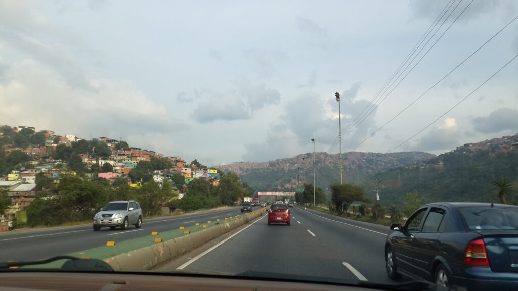 A highway leading toward downtown Caracas, with colorful hillside houses on the left and mountains in the background, seen from inside a moving car.