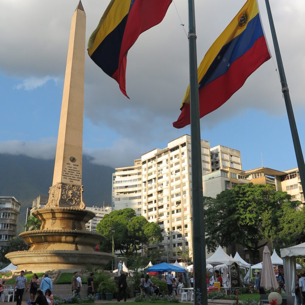 A view of Plaza Francia in Caracas featuring a tall obelisk monument, Venezuelan flags waving in the foreground, surrounding high-rise buildings, trees, and people walking through the plaza.