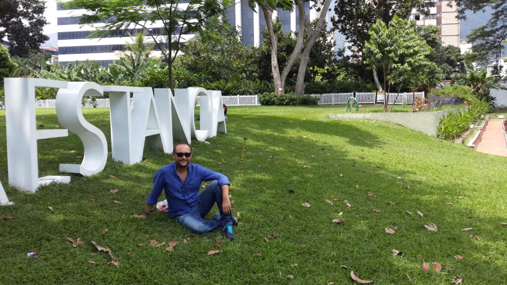 A person sitting on the grass in the gardens of PDVSA Centro de Arte La Estancia in Caracas, beside large white sculptural letters, with lush greenery and tall office buildings in the background.