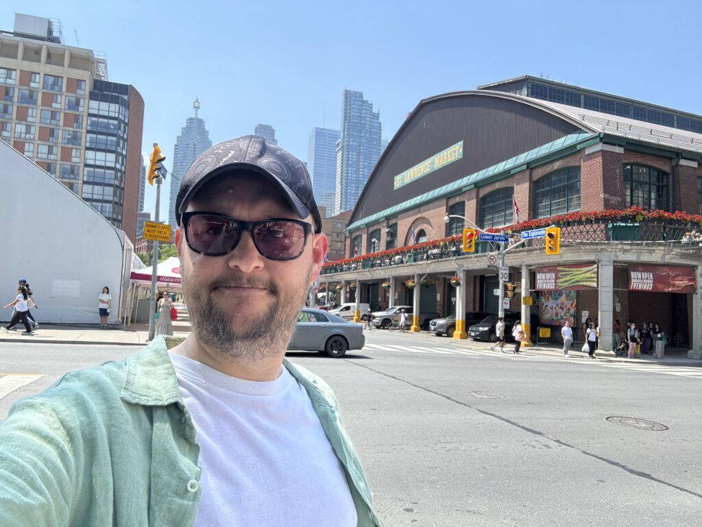 A man wearing sunglasses and a cap taking a selfie in front of Toronto’s St. Lawrence Market (South) on a sunny day, with downtown skyscrapers in the background.