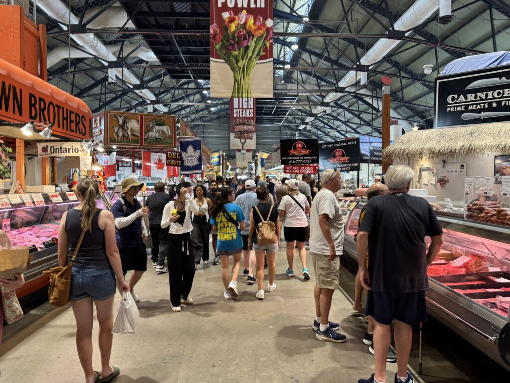 A busy indoor scene at St. Lawrence Market South in Toronto, with people browsing butcher stalls and food vendors under a high industrial-style ceiling.