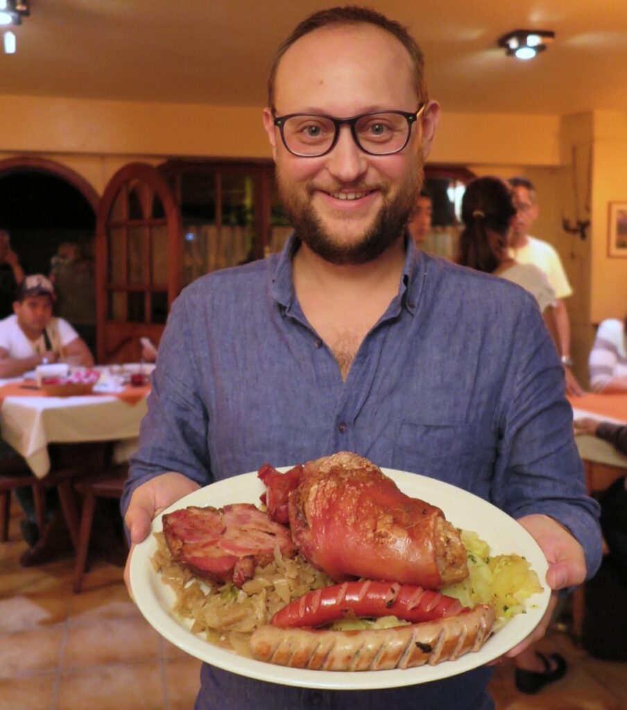 Colonia Tovar Rumbach Restaurant A smiling man in a restaurant holding a large plate of traditional German-style food, including roasted pork knuckle, sausages, and cabbage.