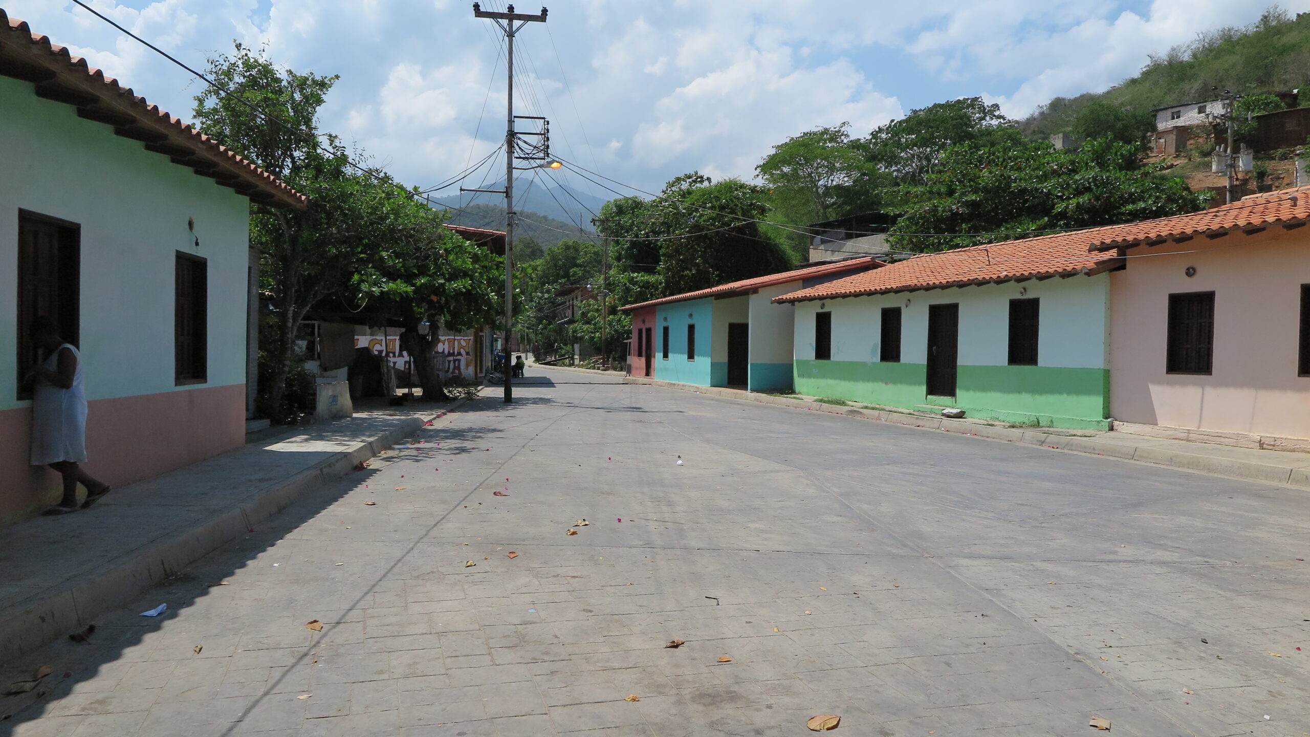 Venezuela'daki Chuao köyünde, parlak bir gökyüzü ve uzaktaki dağ manzarası eşliğinde, sessiz bir taş yolun kenarında sıralanmış renkli kolonyal tarzı evler.