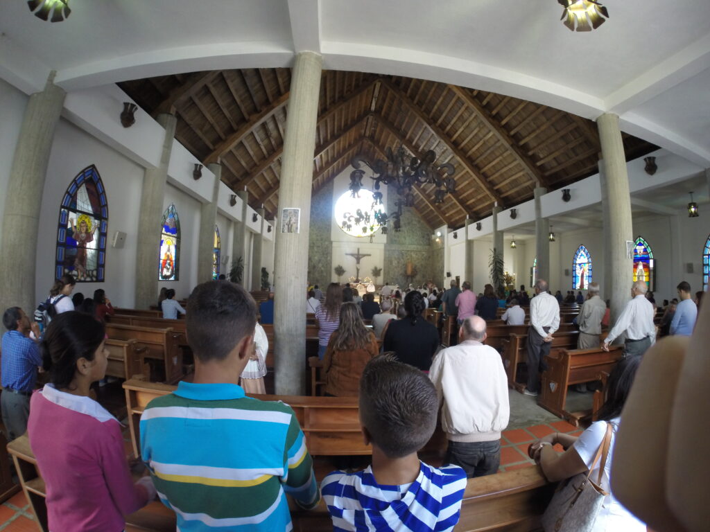 Colonia Tovar - Iglesia San Martín de Tours A wide-angle interior view of San Martín de Tours Church in Colonia Tovar, showing parishioners standing in wooden pews during a religious service. The church features tall columns, a wooden vaulted ceiling, chandeliers, and colorful stained-glass windows along the walls.