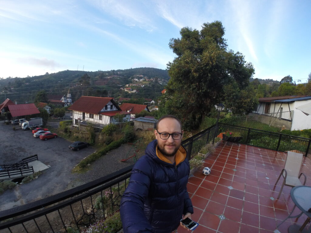Colonia Tovar A person wearing a dark puffer jacket takes a selfie on a tiled terrace overlooking the hillside town of Colonia Tovar, with traditional German-style houses, red roofs, and lush green mountains in the background.