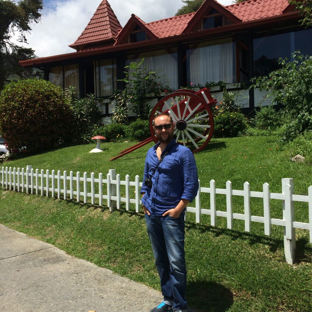 Colonia Tovar A man wearing sunglasses and a blue shirt stands on a sidewalk in front of a traditional German-style house in Colonia Tovar, featuring a red roof, white fence, and a decorative red-and-white wooden wagon wheel on the lawn.
