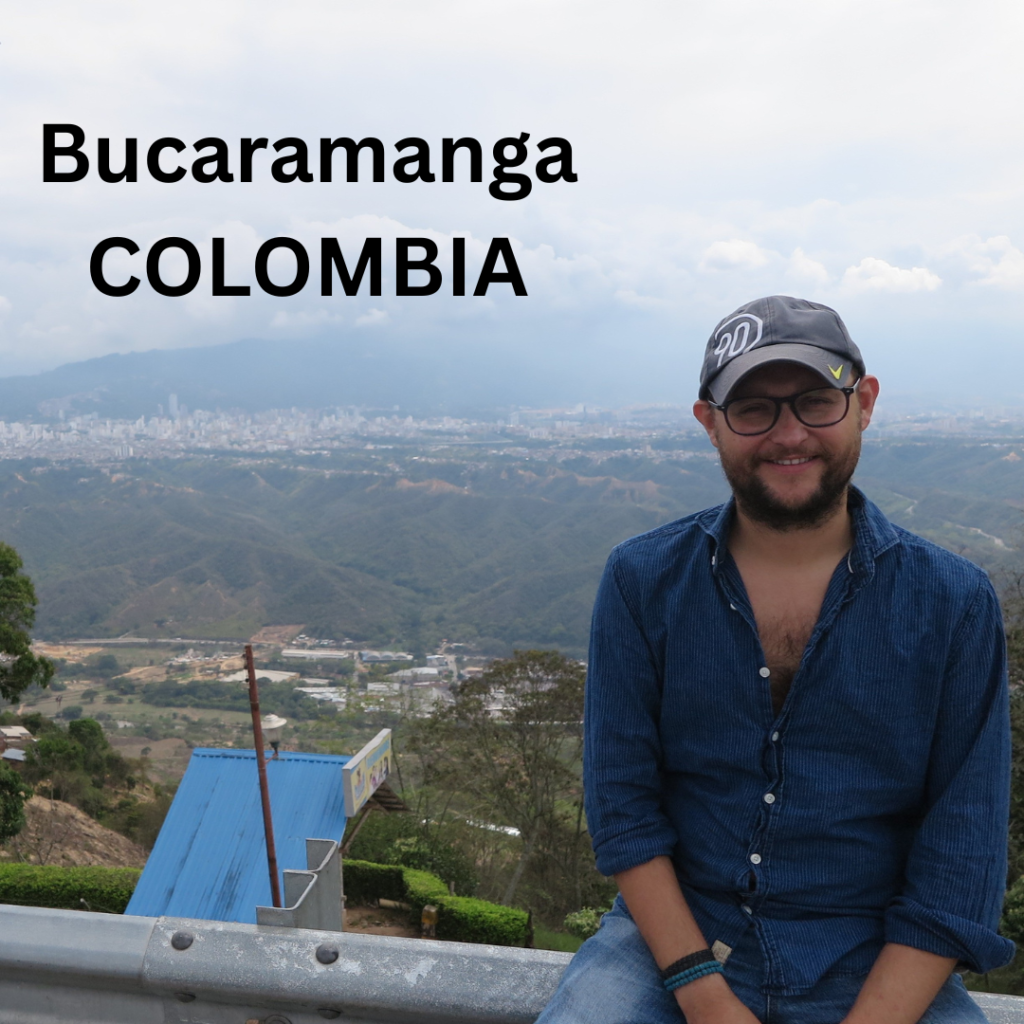 A man smiling in front of a panoramic view of Bucaramanga, Colombia, with green mountains and a cloudy sky in the background.