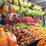 Bucaramanga'daki Plaza Central pazarında, ananas, mango, muz ve narenciye ile dolu canlı bir meyve tezgahının arkasında gülümseyen pembe tişörtlü bir adam ve yerel bir satıcı.