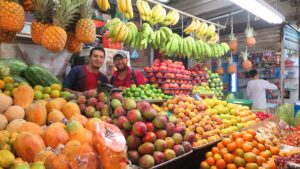 Bucaramanga'daki Plaza Central pazarında, ananas, mango, muz ve narenciye ile dolu canlı bir meyve tezgahının arkasında gülümseyen pembe tişörtlü bir adam ve yerel bir satıcı.