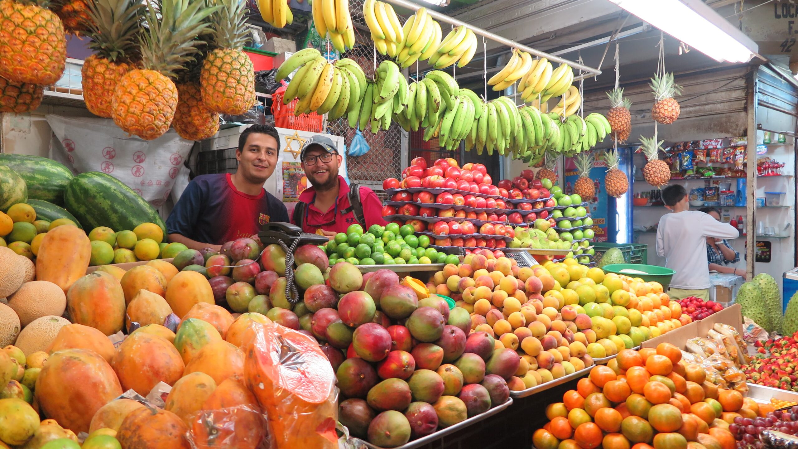Bucaramanga'daki Plaza Central pazarında, ananas, mango, muz ve narenciye ile dolu canlı bir meyve tezgahının arkasında gülümseyen pembe tişörtlü bir adam ve yerel bir satıcı.