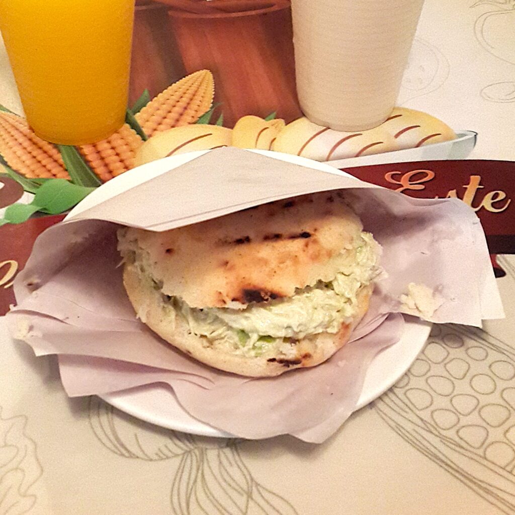 Yanında portakal suyu ile beyaz bir tabakta, kağıt sargı içinde servis edilen; kremsi tavuk ve avokado salatası dolgulu geleneksel Venezuela Arepa Reina Pepiada.
