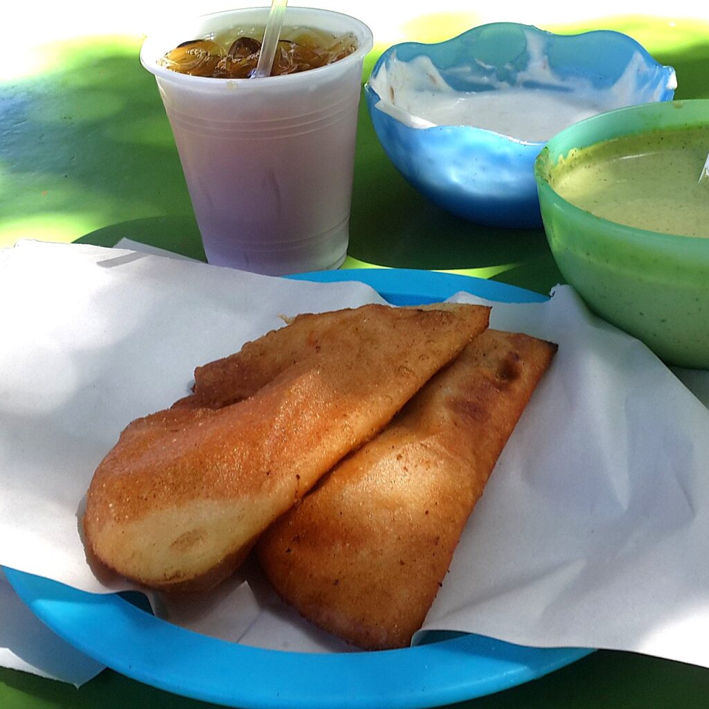 Güneşli bir havada, yeşil masa üzerinde mavi tabakta servis edilen iki adet altın sarısı kızarmış Venezuela empanadası; yanında buzlu içecek ve yeşil sos ile.