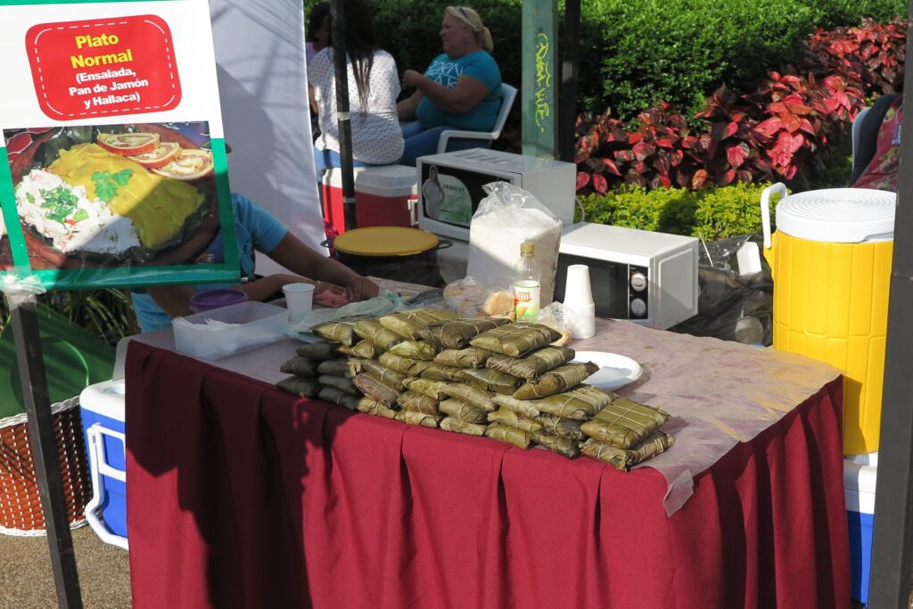 Yeşil muz yapraklarına sarılmış ve iplerle bağlanmış geleneksel Venezuela hallacaları; bir sokak tezgahında "Plato Normal" tabelasının yanında sergileniyor.