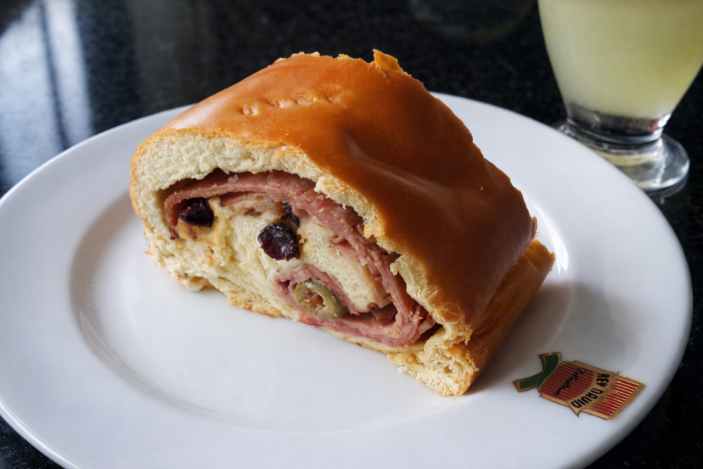 Beyaz tabakta servis edilen bir dilim geleneksel Venezuela Pan de Jamón; altın sarısı tatlı ekmeğin içindeki jambon, zeytin ve kuru üzüm dolgusu görülüyor.