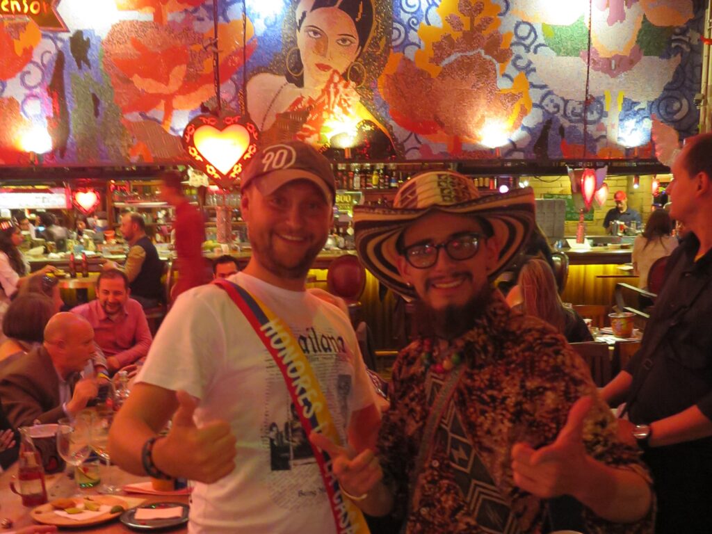 Two men smiling and posing for a photo inside Andrés Carne de Res Bogota, with one wearing a traditional Colombian Sombrero Vueltiao and the other a celebratory sash.