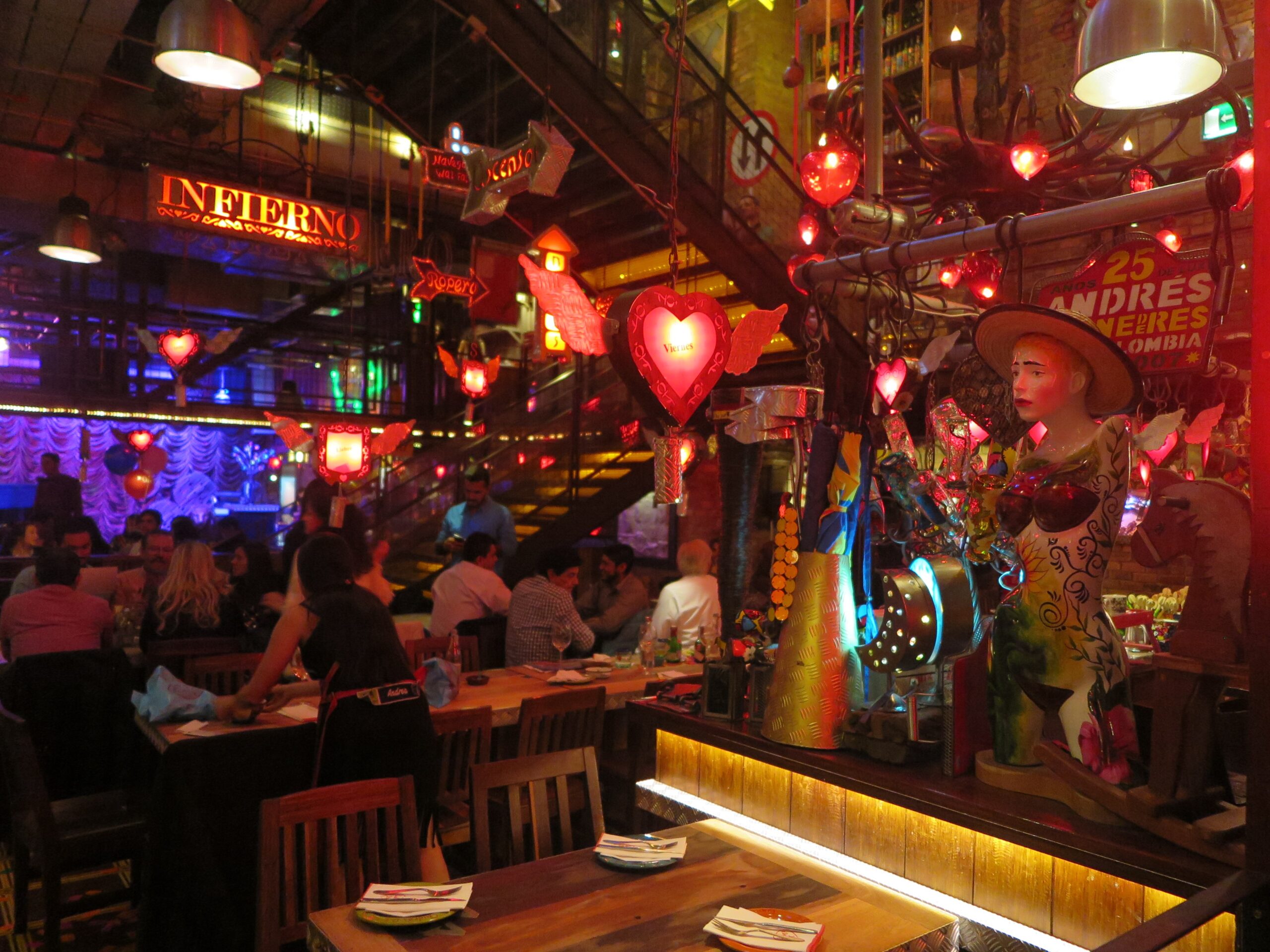 Interior of Andrés Carne de Res (Andrés D.C.) in Bogotá, filled with glowing red heart-shaped neon lights, eclectic décor, a colorful statue, and guests dining at wooden tables.
