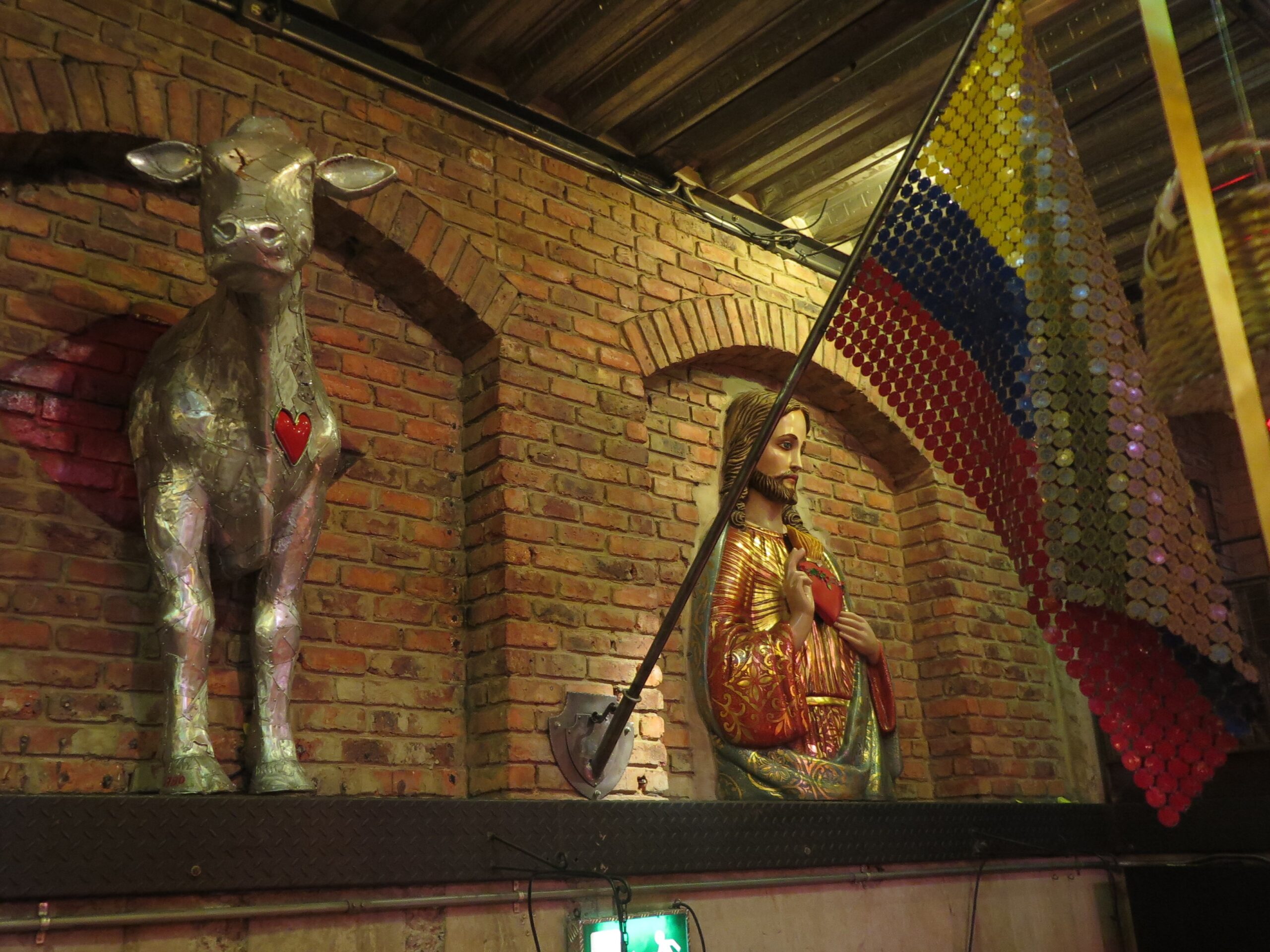 Quirky interior decor at Andrés Carne de Res in Bogota featuring a silver cow statue and a religious figure with a red heart on a brick wall.