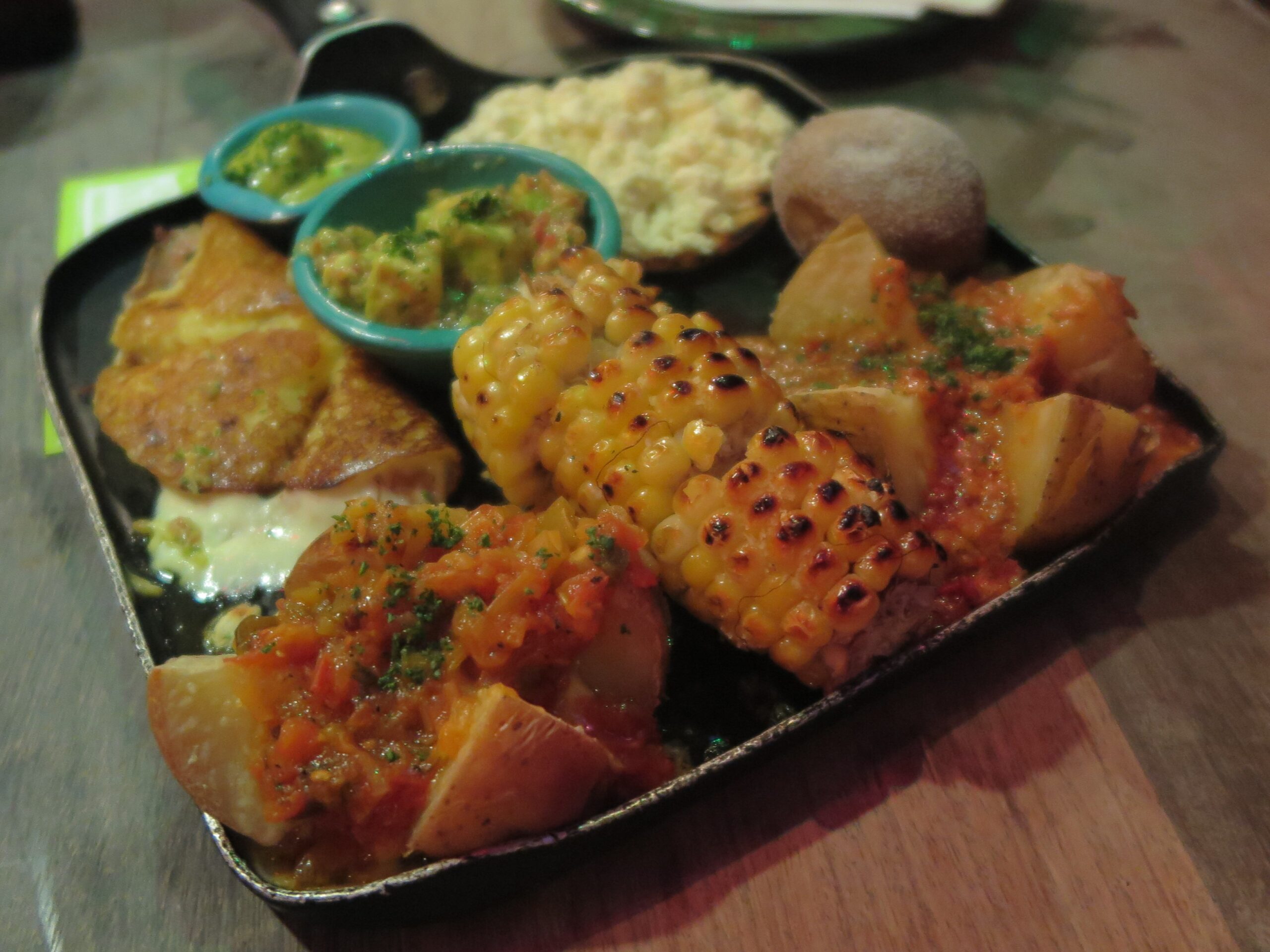 Close-up of a Colombian appetizer platter at Andrés Carne de Res featuring grilled corn on the cob, papas criollas with hogao sauce, an arepa, and bowls of guacamole and salsa.
