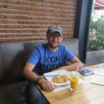 A man in a blue t-shirt and grey cap sitting at a wooden table inside Crepes & Waffles in Bogota. He has a plate with a savory crepe and a glass of fresh orange juice in front of him. A brick wall is behind him.