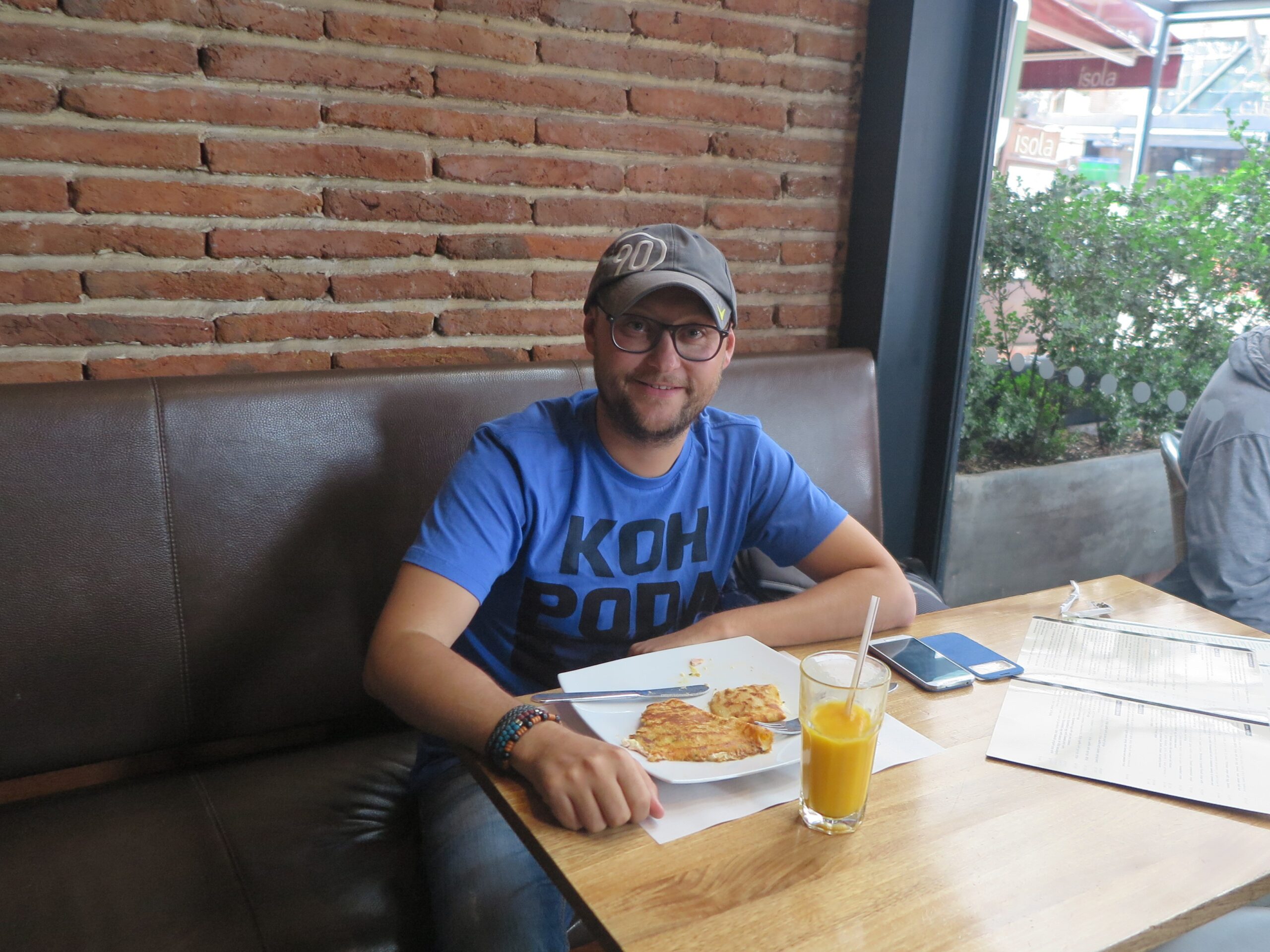 A man in a blue t-shirt and grey cap sitting at a wooden table inside Crepes & Waffles in Bogota. He has a plate with a savory crepe and a glass of fresh orange juice in front of him. A brick wall is behind him.