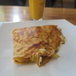A close-up shot of a golden-brown savory crepe filled with melted cheese and ham on a white square plate. A glass of orange juice is blurred in the background on a wooden table.