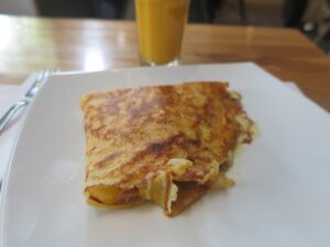 A close-up shot of a golden-brown savory crepe filled with melted cheese and ham on a white square plate. A glass of orange juice is blurred in the background on a wooden table.