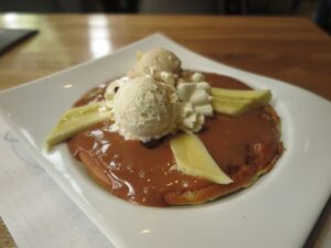 A dessert waffle on a white square plate, smothered in rich caramel sauce (arequipe) and topped with slices of banana, two scoops of ice cream, and whipped cream.