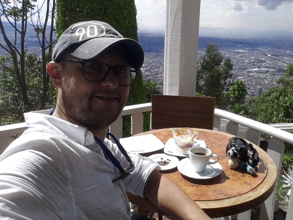 Bogota Monserrate Mountain 1 A man wearing a grey cap and glasses sitting at a cafe table on Mount Monserrate, Bogota, with a cup of coffee and a dessert. The vast city of Bogota is visible in the background under a cloudy sky.
