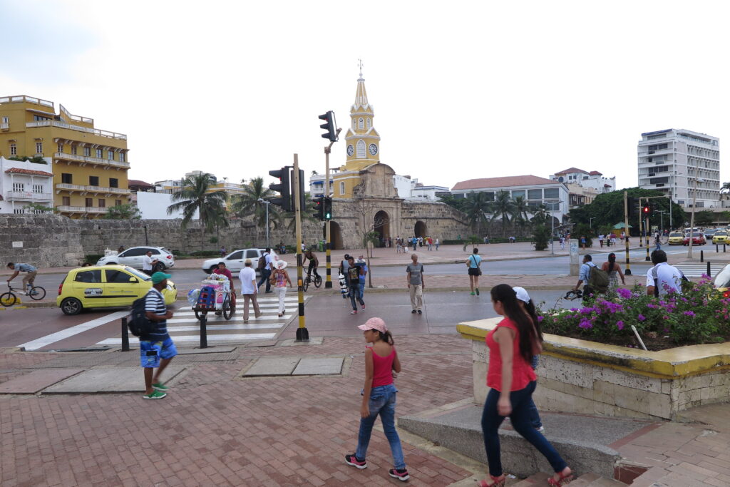 A busy street scene in Cartagena, Colombia, featuring the iconic yellow Clock Tower (Torre del Reloj) and people walking near the historic stone walls.