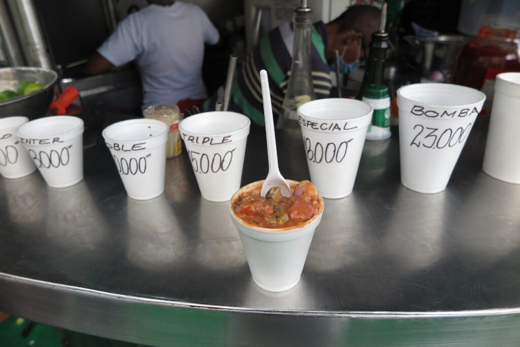 A close-up of a fresh shrimp cocktail in a white cup, placed on a metal counter with other cups showing prices like 'Bomba' and 'Triple' at a street food stall in Cartagena