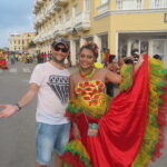 A traveler posing with a local woman in a colorful traditional dress during Cartagena Independence Day celebrations, with a lively street parade and historic yellow colonial buildings in the background.