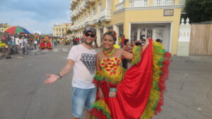 A traveler posing with a local woman in a colorful traditional dress during Cartagena Independence Day celebrations, with a lively street parade and historic yellow colonial buildings in the background.