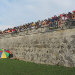 A long shot of the historic stone walls of Cartagena, Colombia, during Cartagena Independence Day. A dense line of spectators sits along the very top edge of the high wall, overlooking a green field.