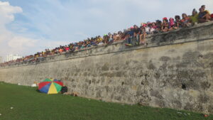 A long shot of the historic stone walls of Cartagena, Colombia, during Cartagena Independence Day. A dense line of spectators sits along the very top edge of the high wall, overlooking a green field.
