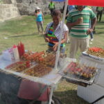 A street food vendor in Cartagena, Colombia, grilling various meat and potato skewers on a small outdoor grill during Independence Day. Colorful umbrellas provide shade for the stall.