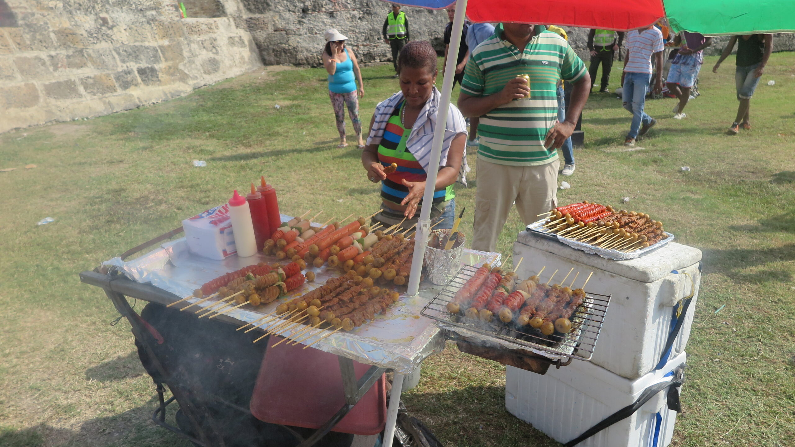 A street food vendor in Cartagena, Colombia, grilling various meat and potato skewers on a small outdoor grill during Independence Day. Colorful umbrellas provide shade for the stall.