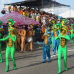 Colorfully dressed performers in snake-themed costumes marching during Cartagena Independence Day parade, with musicians, spectators, and packed grandstands in the background.