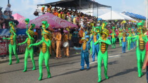 Colorfully dressed performers in snake-themed costumes marching during Cartagena Independence Day parade, with musicians, spectators, and packed grandstands in the background.