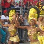 Dancers wearing gold costumes and tall decorative headpieces perform during Cartagena Independence Day parade, with confetti in the air and a cheering crowd behind barriers.