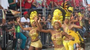Dancers wearing gold costumes and tall decorative headpieces perform during Cartagena Independence Day parade, with confetti in the air and a cheering crowd behind barriers.