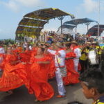 Dancers in bright orange traditional dresses and musicians parade through the streets of during Cartagena Independence Day celebrations, with crowds watching and a seaside stage in the background.