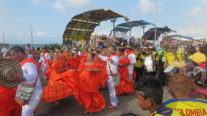 Dancers in bright orange traditional dresses and musicians parade through the streets of during Cartagena Independence Day celebrations, with crowds watching and a seaside stage in the background.